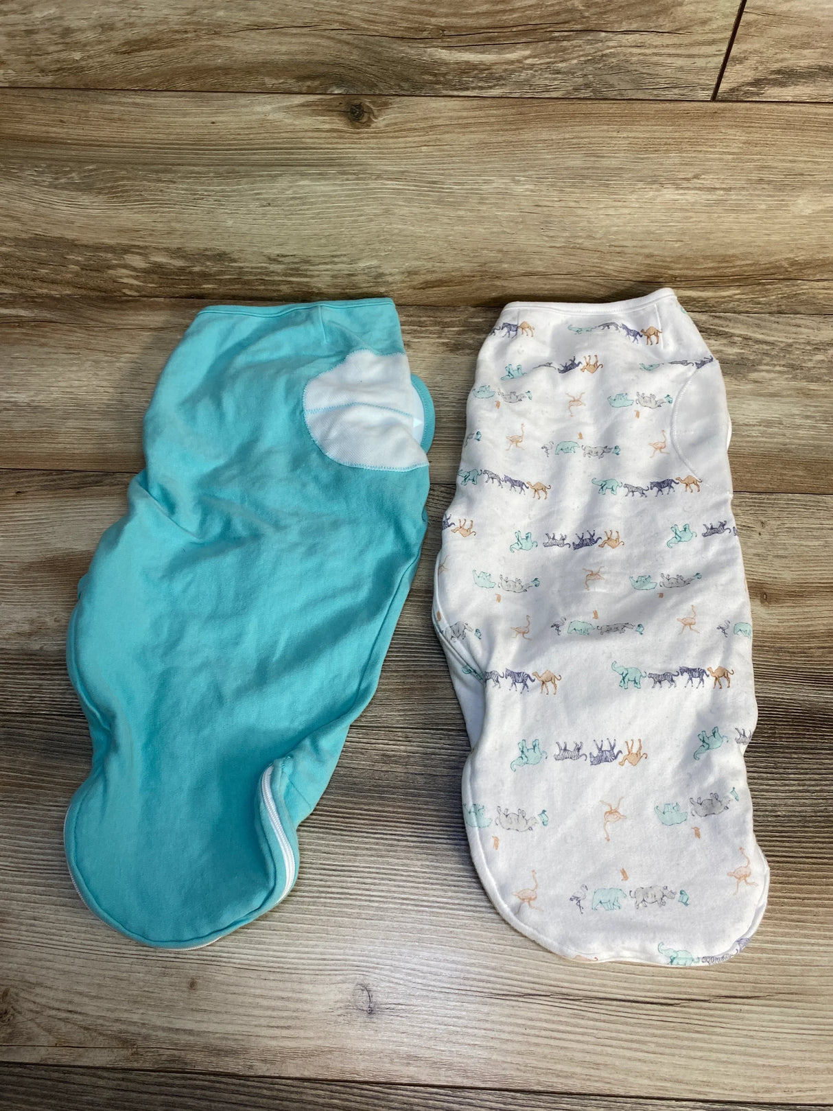 Flat lay of Aden + Anais 2-pack newborn swaddle wraps 0-3M showing one solid teal wrap and one white animal-print wrap on a wood floor