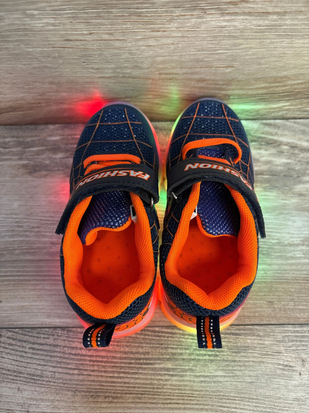 Top-down view of navy mesh toddler sneakers with bright orange lining and Velcro strap, multicolor light-up soles on wood floor