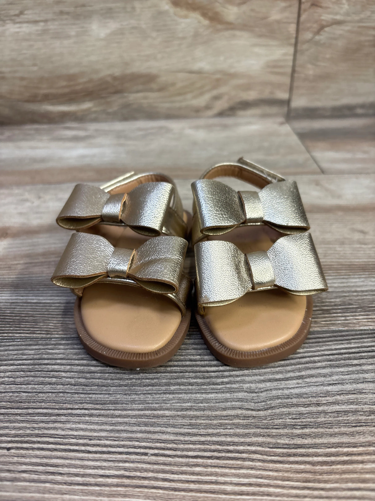 Front view of Cat & Jack toddler gold Ruby bow sandals showcasing two metallic bows on double straps and a tan cushioned footbed on a wood-grain floor
