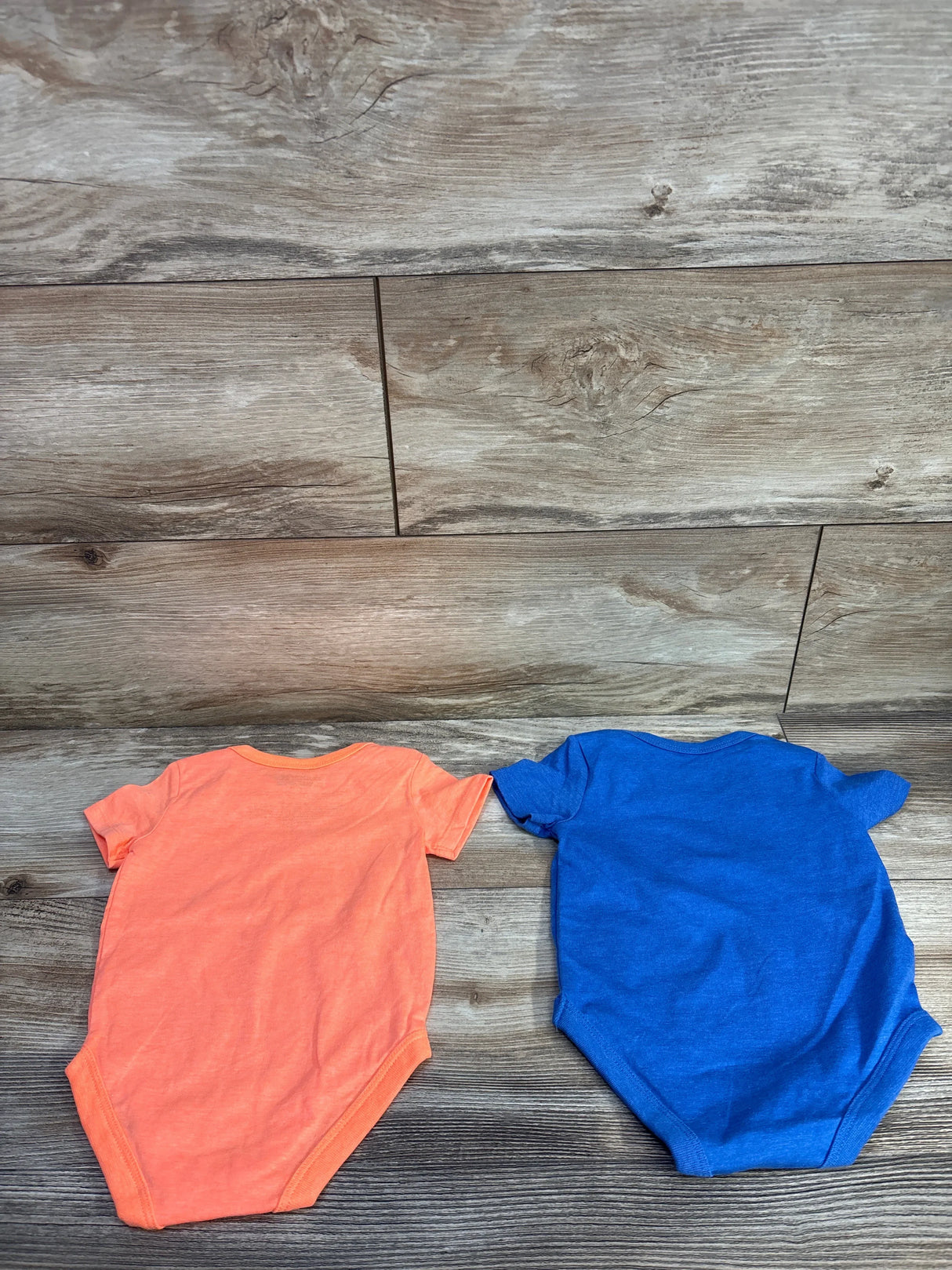 Flat lay of two Hurley short-sleeve baby bodysuits showing plain orange and blue backs on wood floor
