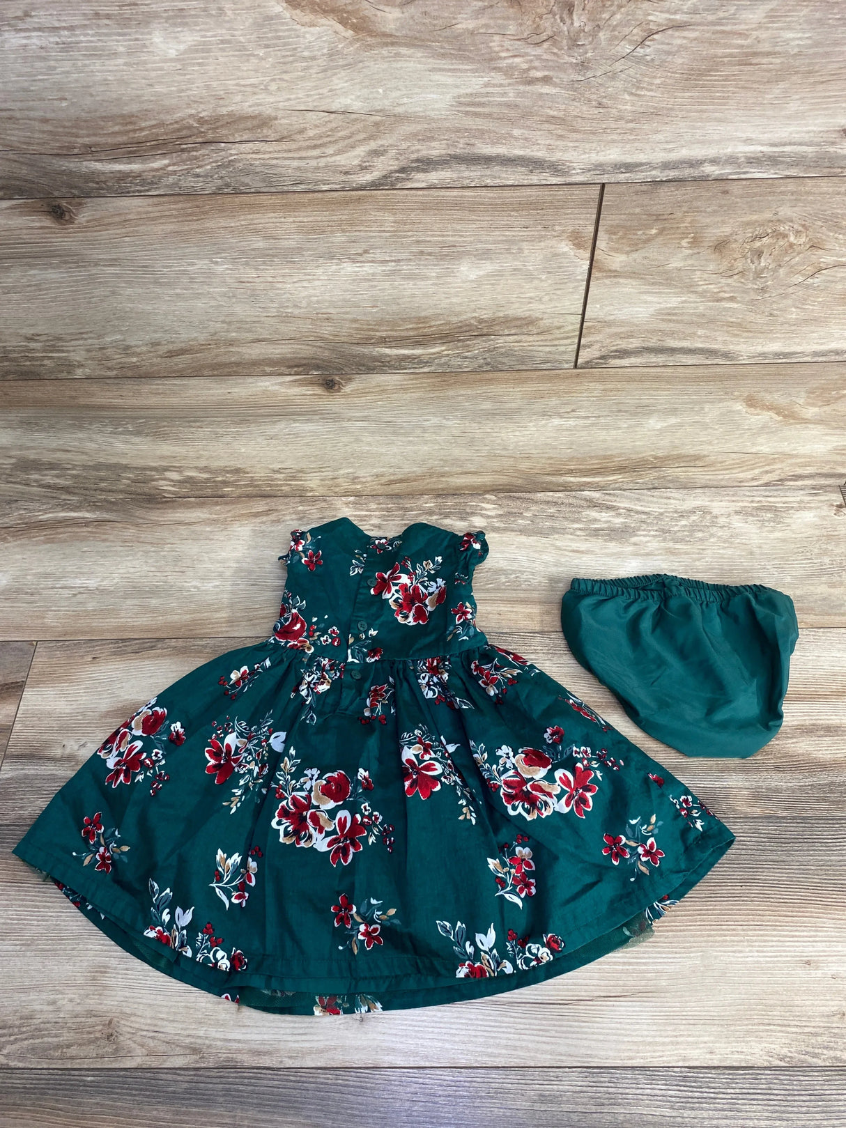 Green infant dress with red and white floral print and matching green diaper cover on wooden floor