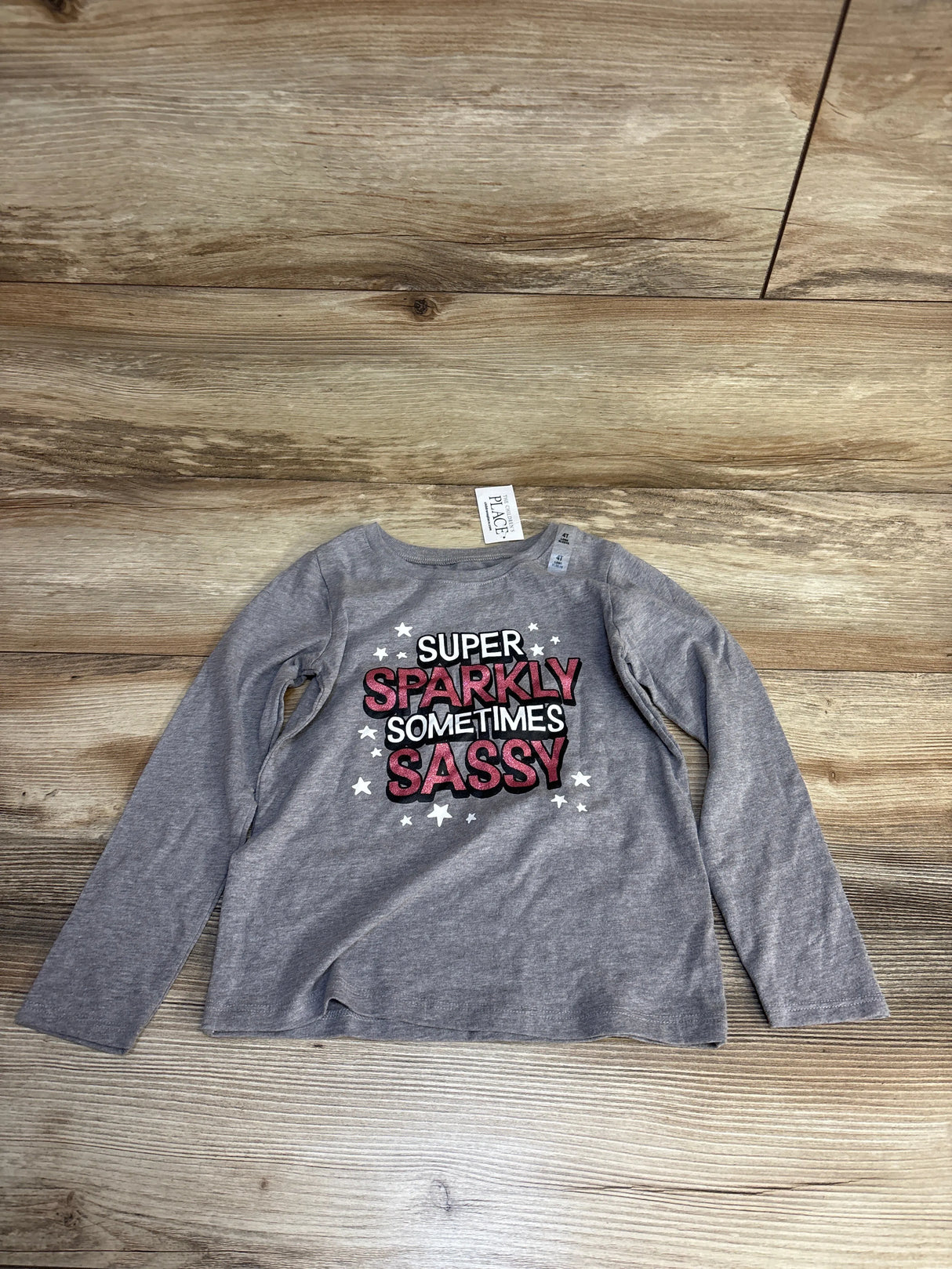 Gray kids long sleeve shirt with 'Super Sparkly Sometimes Sassy' graphic and stars, laid on wood floor