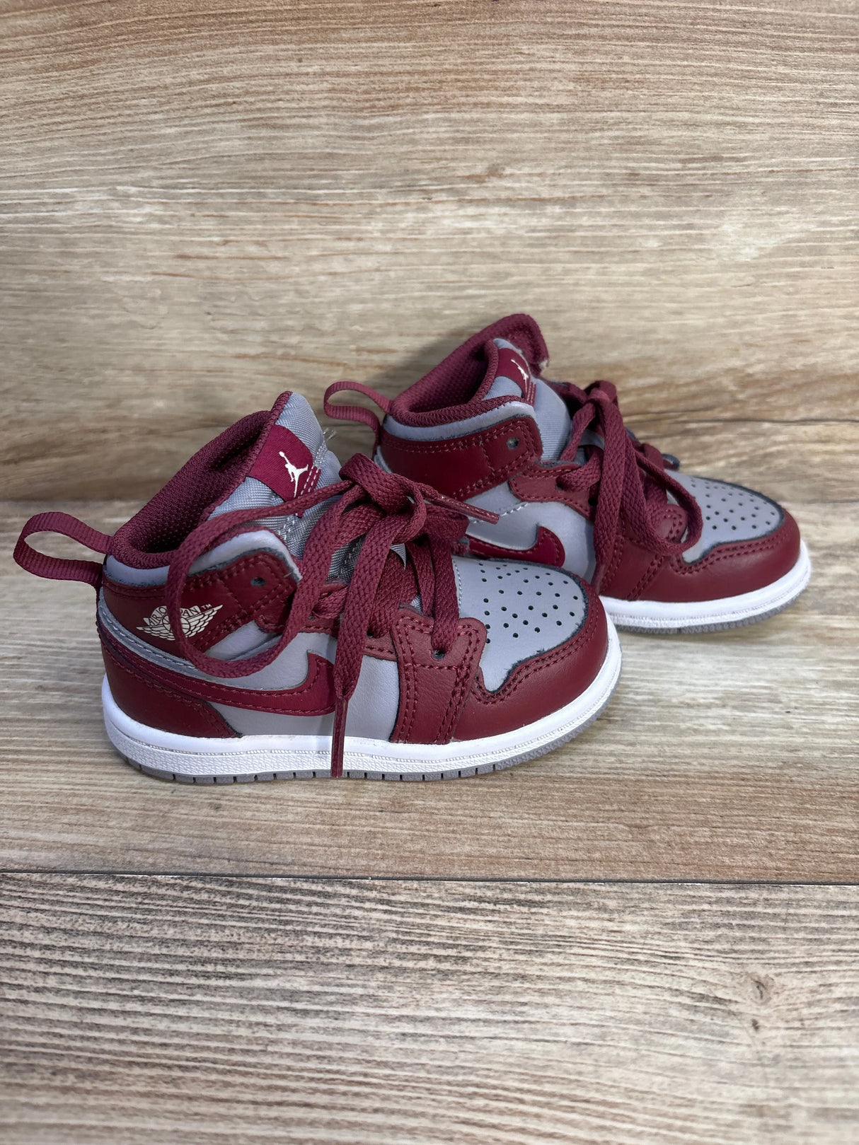 Side view of Air Jordan 1 Mid TD Cherrywood Red toddler sneakers size 6C showing maroon leather overlays, gray panels, laces, and white midsole on wood background