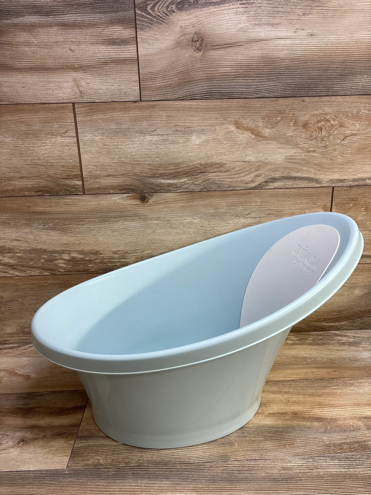 Angled side view of BEABA Shnuggle baby tub, pale blue, showing reclined shape and white foam backrest on wood background