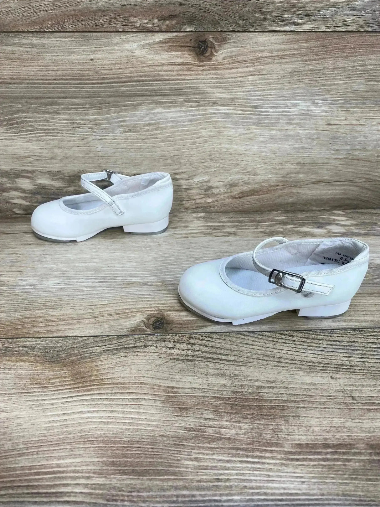 Angled view of white child tap buckle shoe showing buckle strap, low block heel, and rounded toe on light wood background