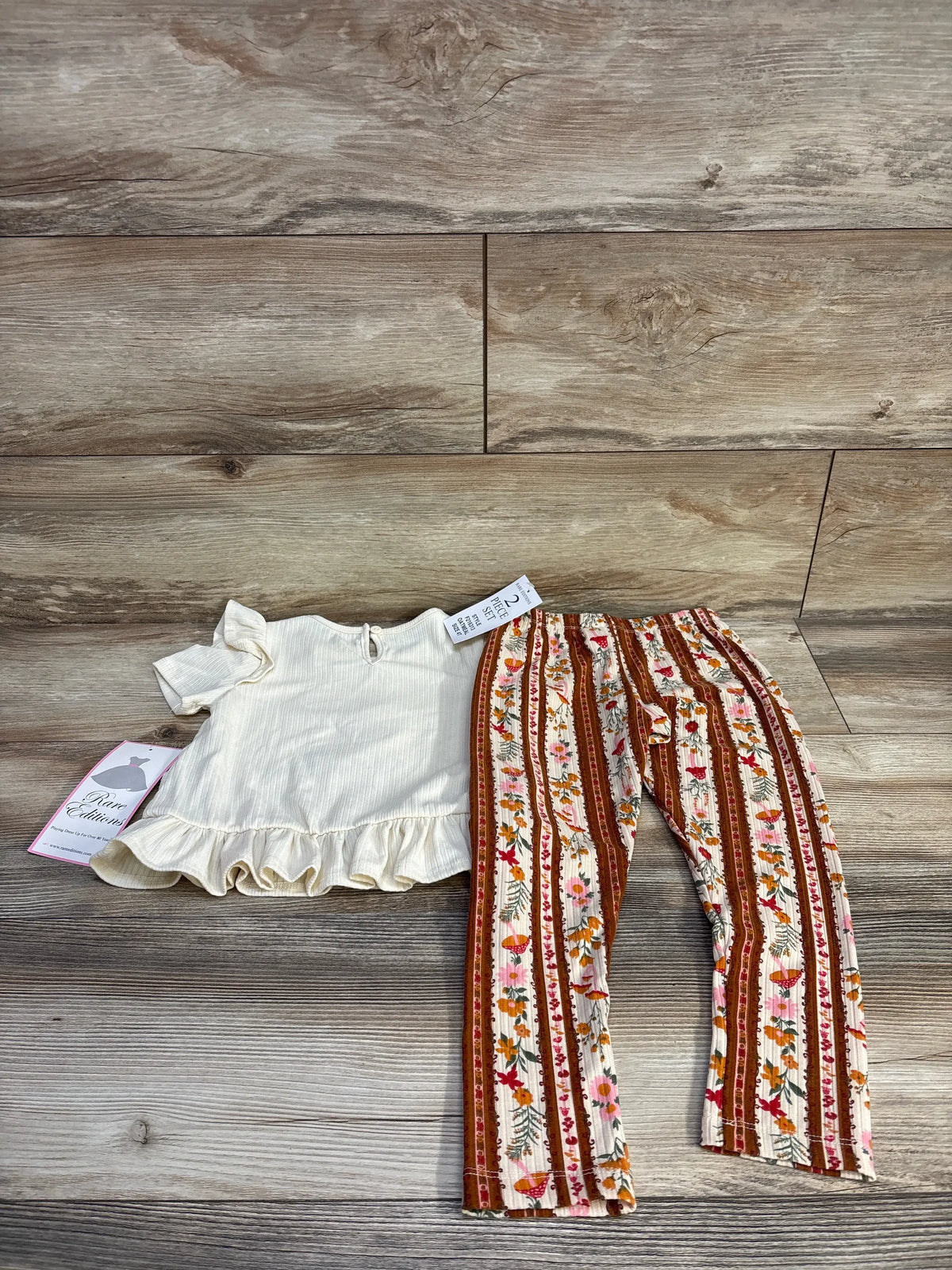 Overhead view of Rare Editions toddler two-piece outfit with cream ribbed peplum shirt and floral striped pants on wood
