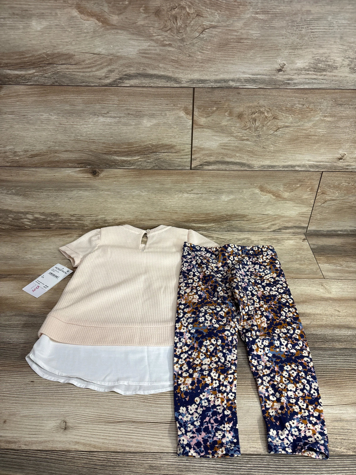 Flat lay of 2-piece toddler outfit showing cream top with white layered hem and keyhole back; navy floral leggings on wood floor
