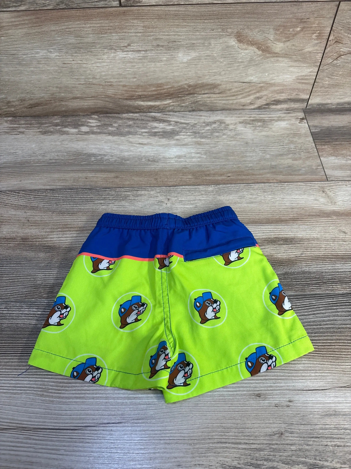 Flat lay of Buc-ees toddler swim trunks, back view showing blue elastic waistband and neon green fabric with repeating beaver logo on wood floor