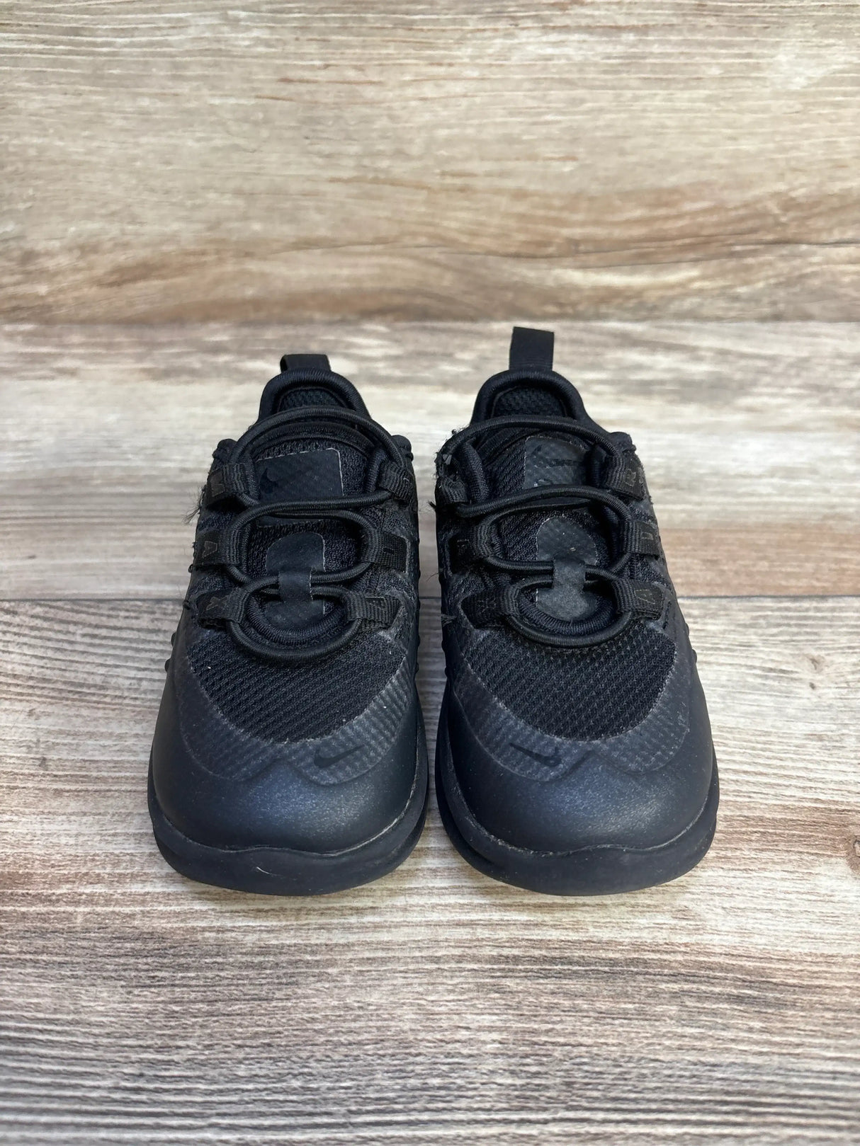 Front view of toddler Nike Air Max Axis black sneakers showcasing mesh upper, bungee laces and heel pull tabs on wood background