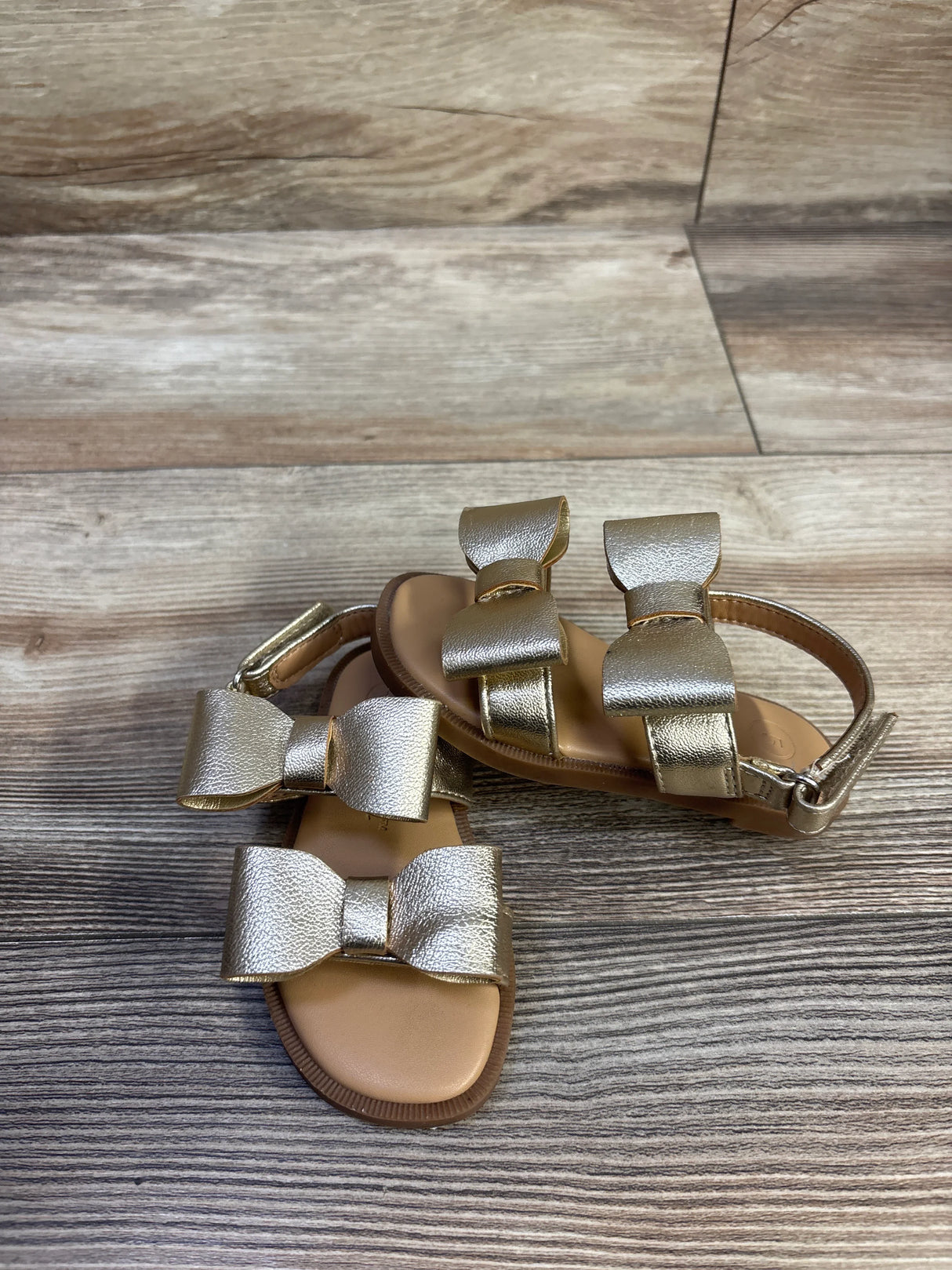 Angled top view of Cat and Jack toddler bow sandals showcasing metallic gold bows and ankle strap on wood floor background