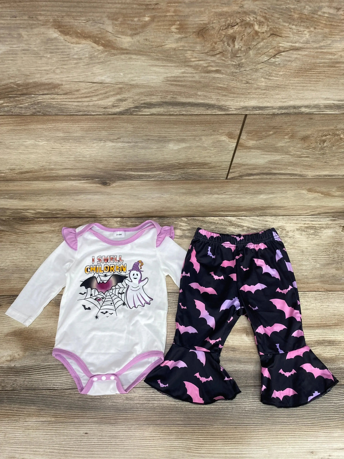 Flat lay of white baby bodysuit with I Smell Children ghost and matching black ruffle pants with pink and purple bats on wood