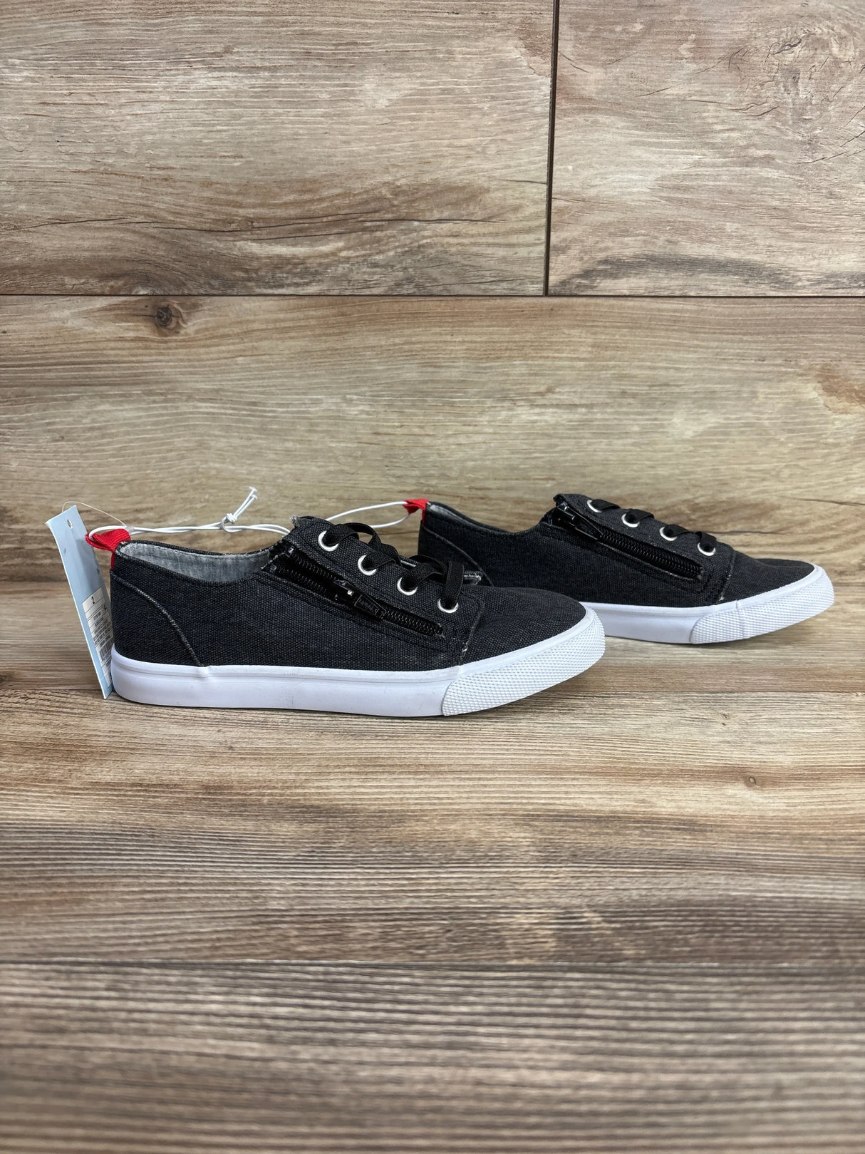 Pair of black kids canvas sneakers with white soles, lateral zippers, and red heel tabs on wood floor
