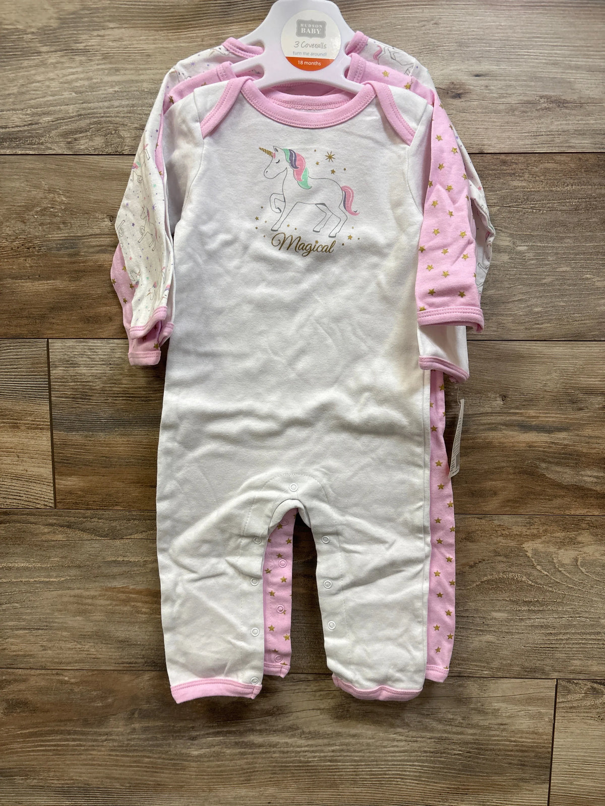 Front view of Hudson Baby three pack coveralls in 18 month size showcasing white and pink unicorn and star patterns on wood floor