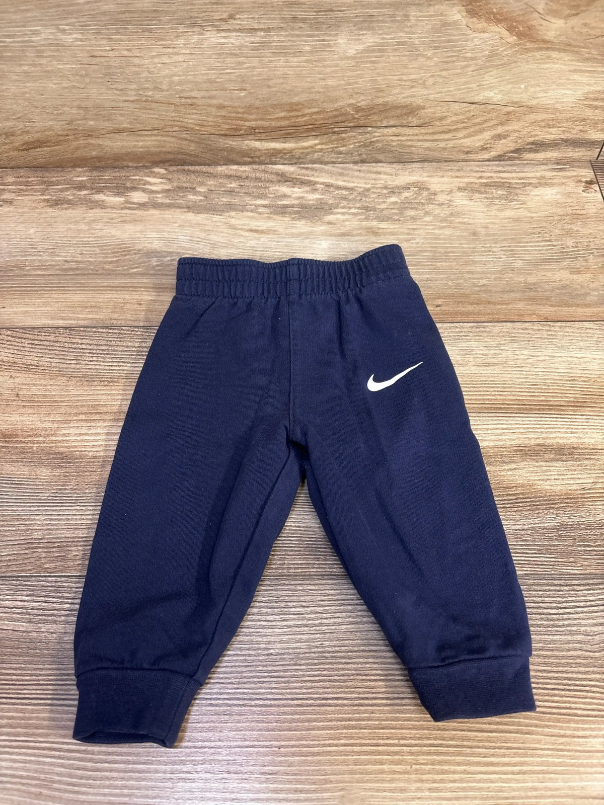 Flat lay of navy baby Nike pull-on joggers size 6M showing elastic waistband, ribbed cuffs and white Swoosh on wood floor