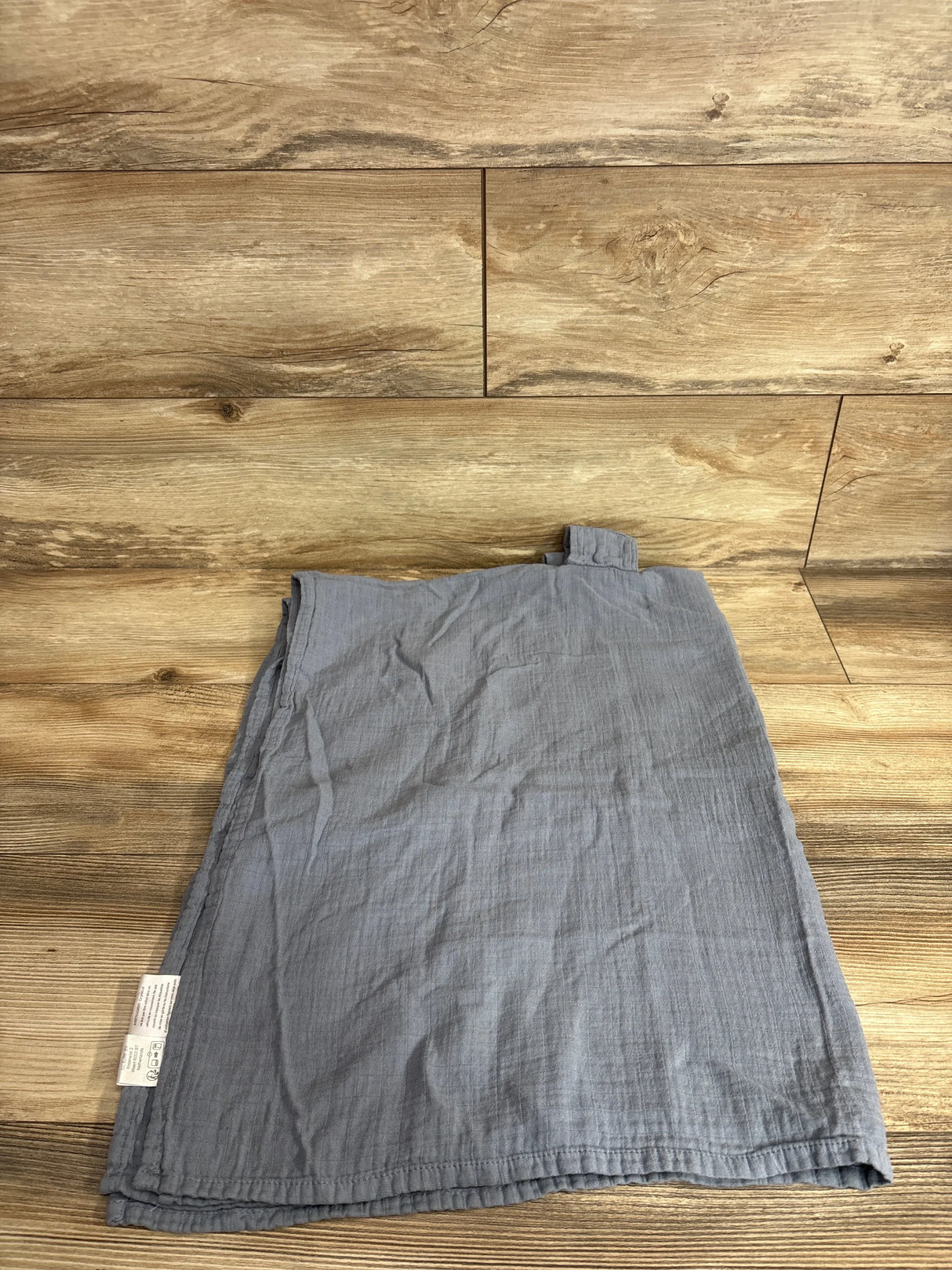 Flat lay of Comfy Cubs muslin car seat cover in blue showing crinkled muslin texture and top attachment tab on wood floor background