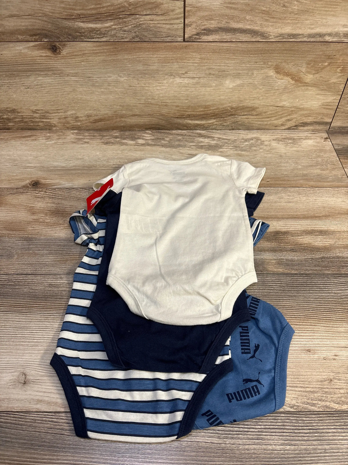 Top-down view of 4-pack baby bodysuits (3-6M) stacked: white, navy, blue striped, blue Puma logo print on wooden background