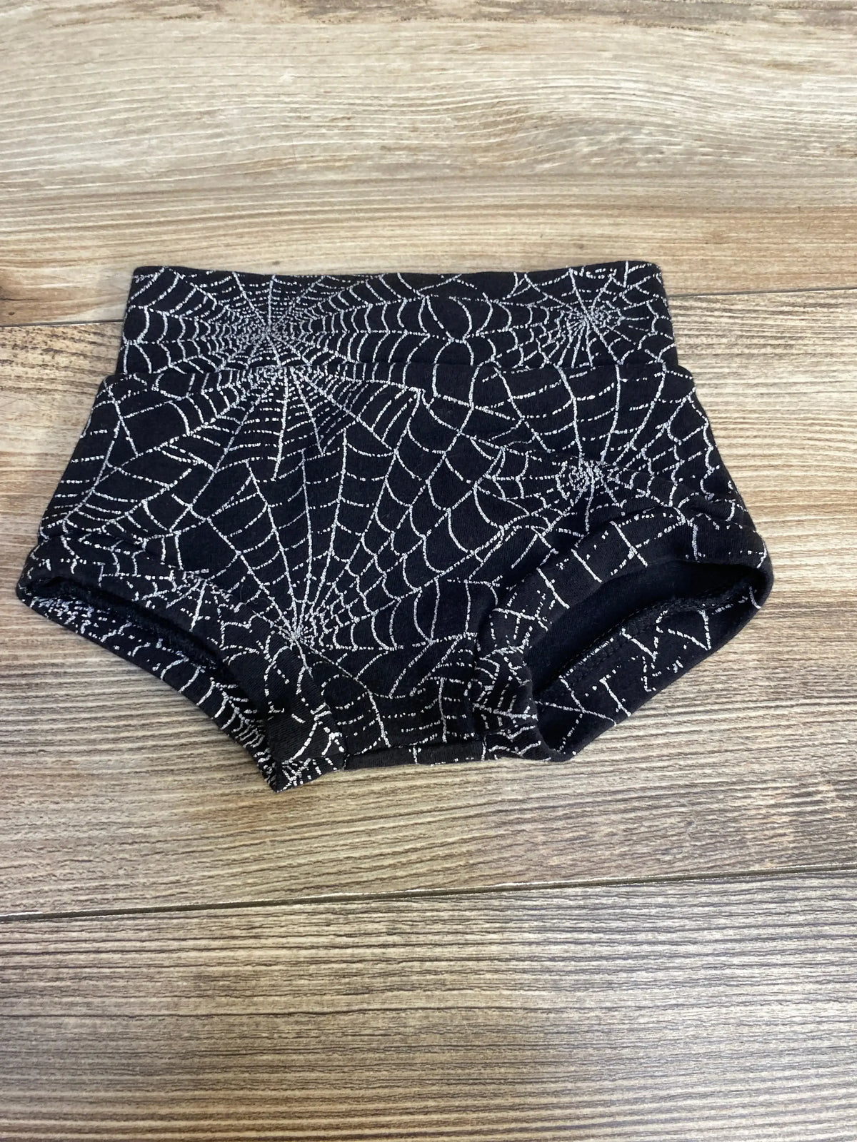 Top-down of black baby bloomers with white spiderweb print, high-rise waistband and elastic leg openings on wooden floor