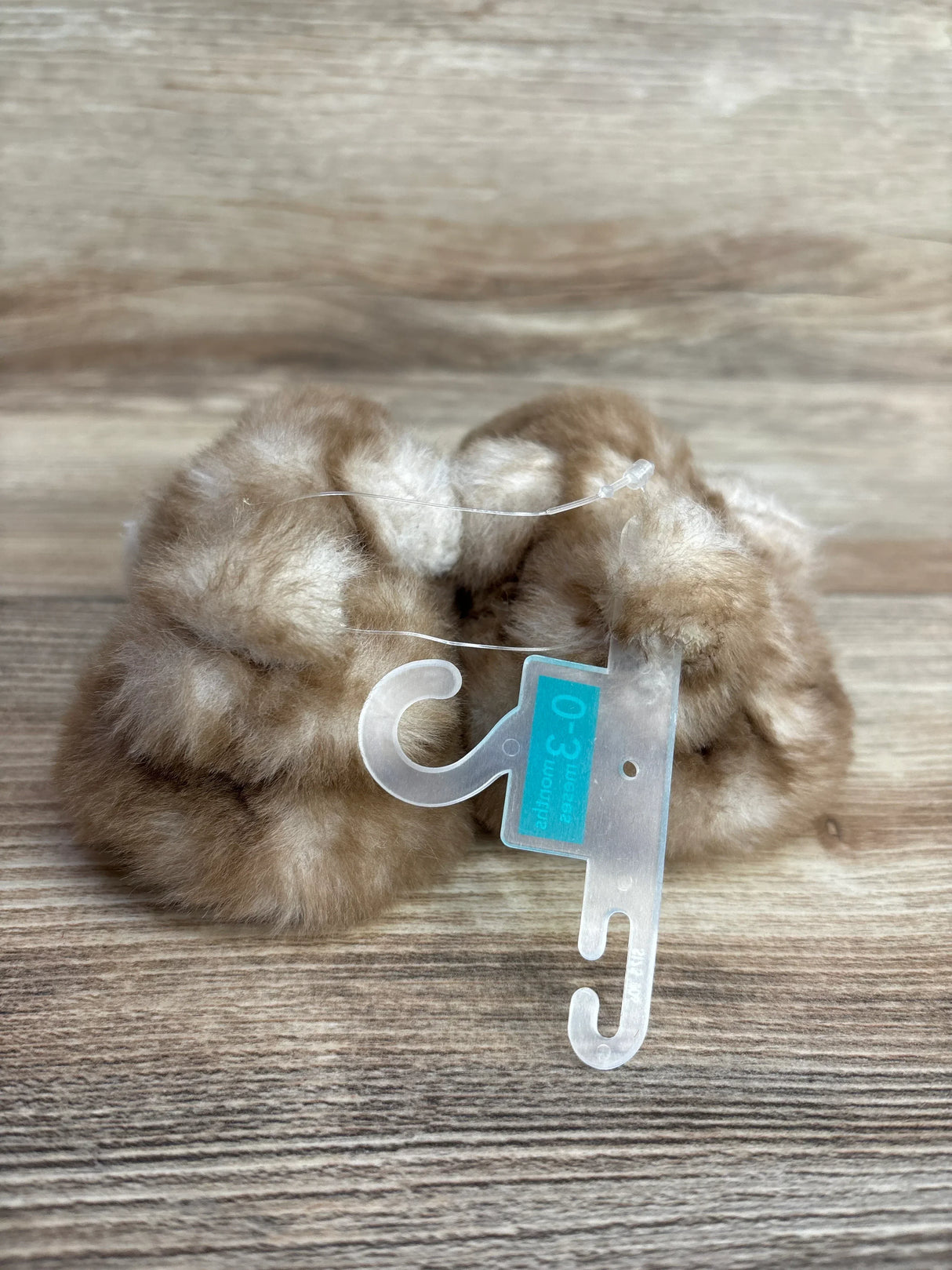 Pair of soft, beige, faux fur baby booties with size 0-3 months tag on wooden surface