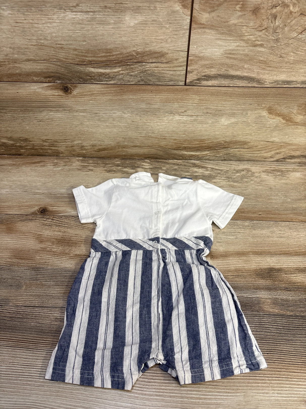Flat lay of white baby romper with navy vertical striped suspender shorts and back buttons on wooden floor