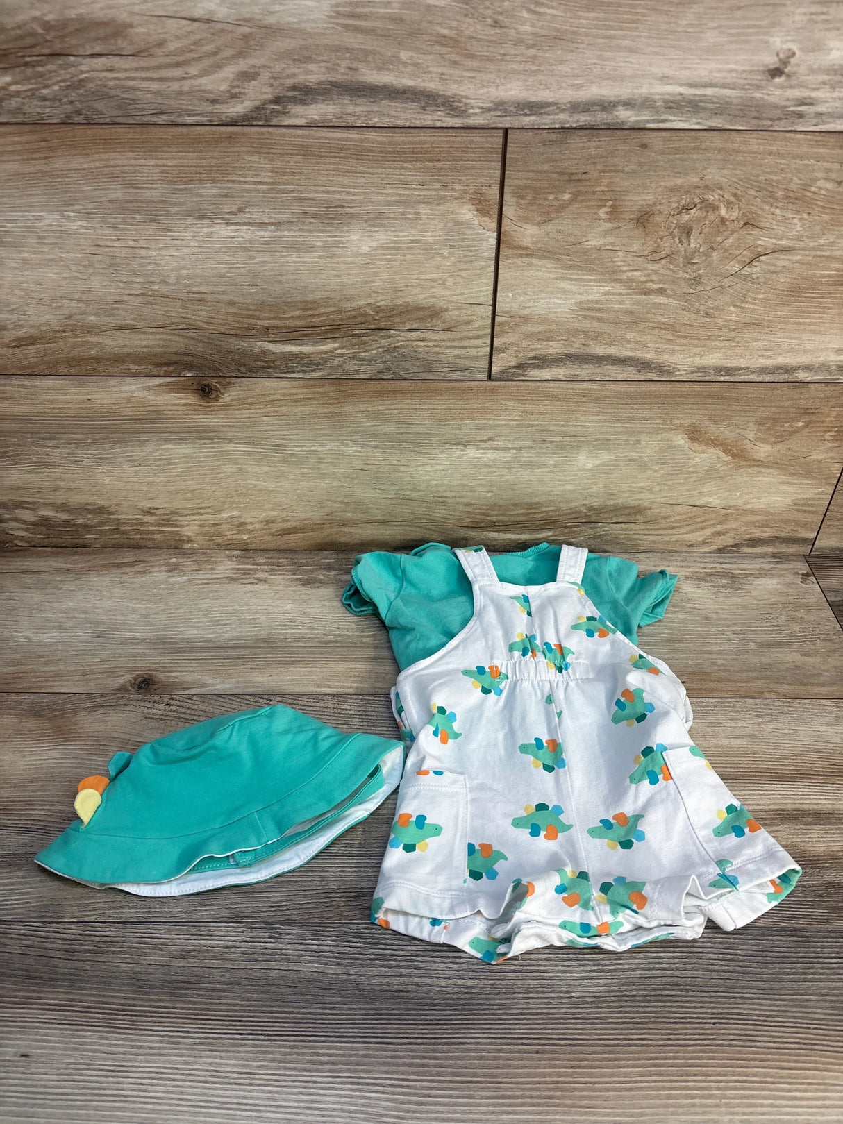 Flat lay of Mayoral turquoise dinosaur dungaree set 3-6M: white romper with dinosaur print, teal tee and hat on wood