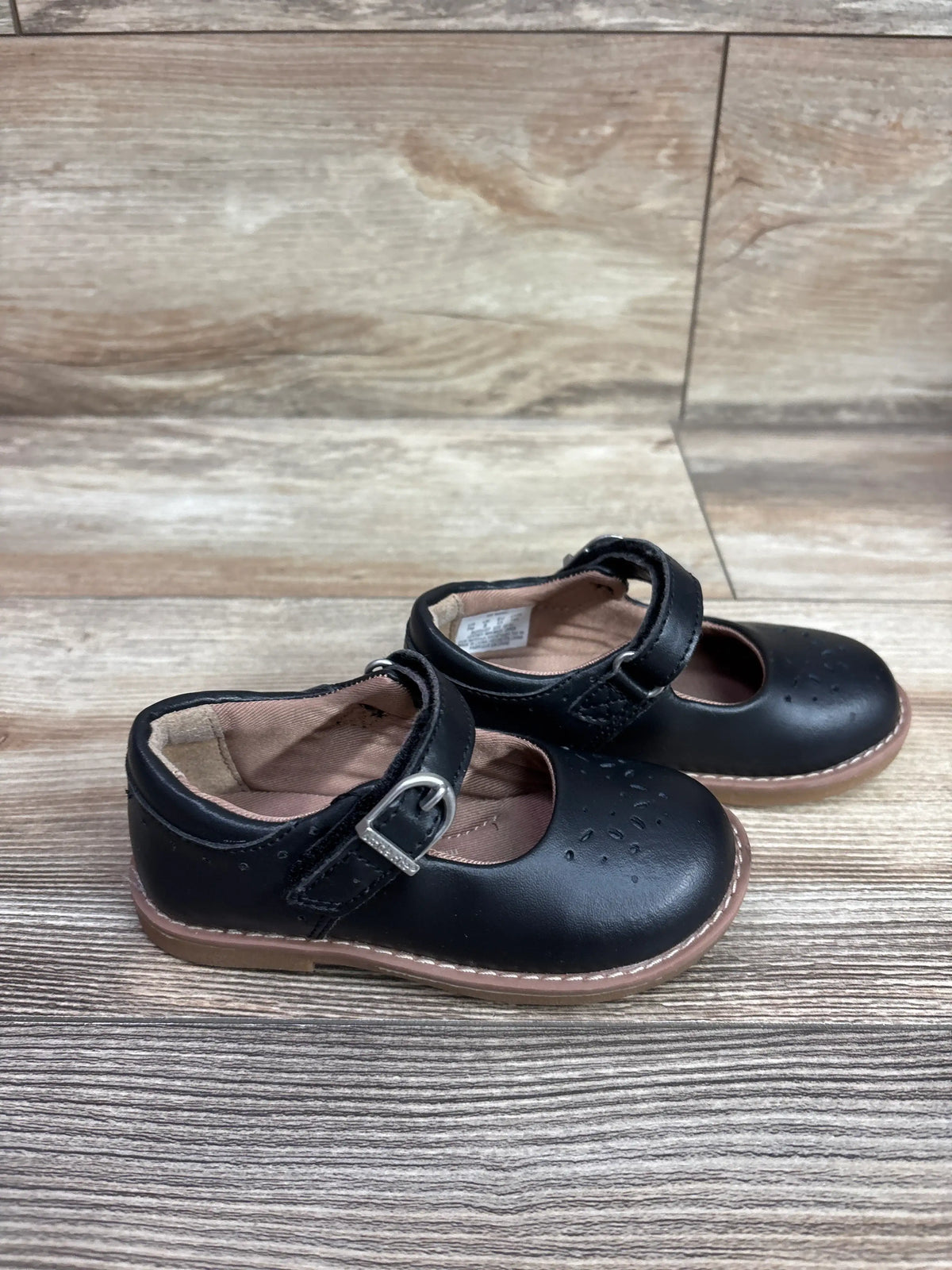 Angled view of black Stride Rite Mara Mary Jane flats with buckle straps and perforated toes on a wood-grain floor