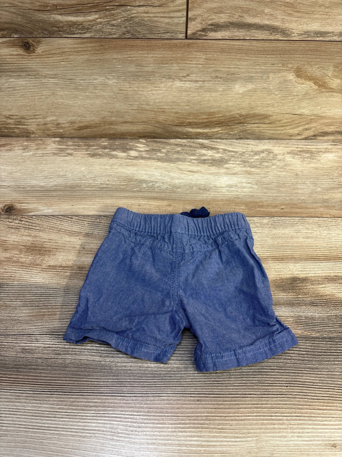 Flat-lay of blue chambray baby shorts (3-6 months) showing elastic waistband and rolled cuffs on wood background