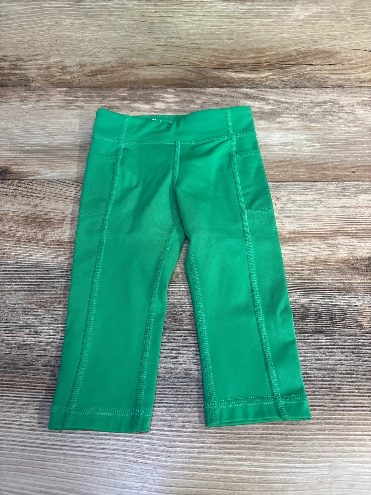 Top down view of green toddler active leggings (3T) showing wide elastic waistband, reinforced flat-lock seams and cropped length on wood background