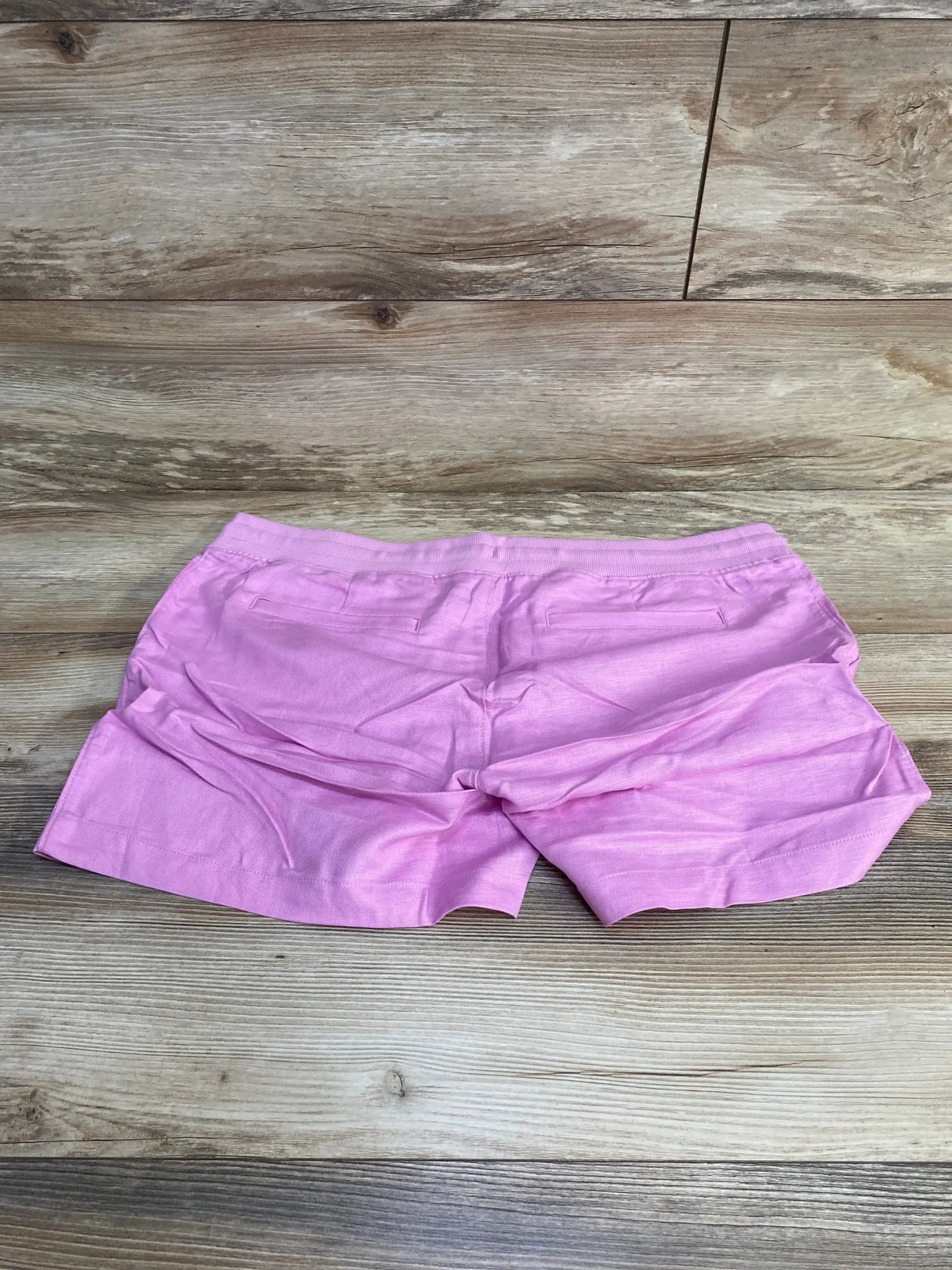Back view of Gap pink maternity linen shorts showing elastic waistband, faux back welt pockets and cuffed hem on wood floor