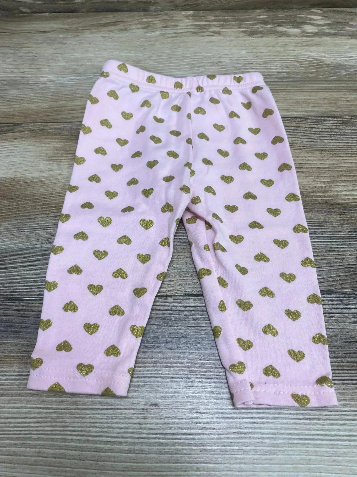 Flat lay of pink baby leggings (3-6 months) showcasing gold glitter heart print and elastic waistband on wood background