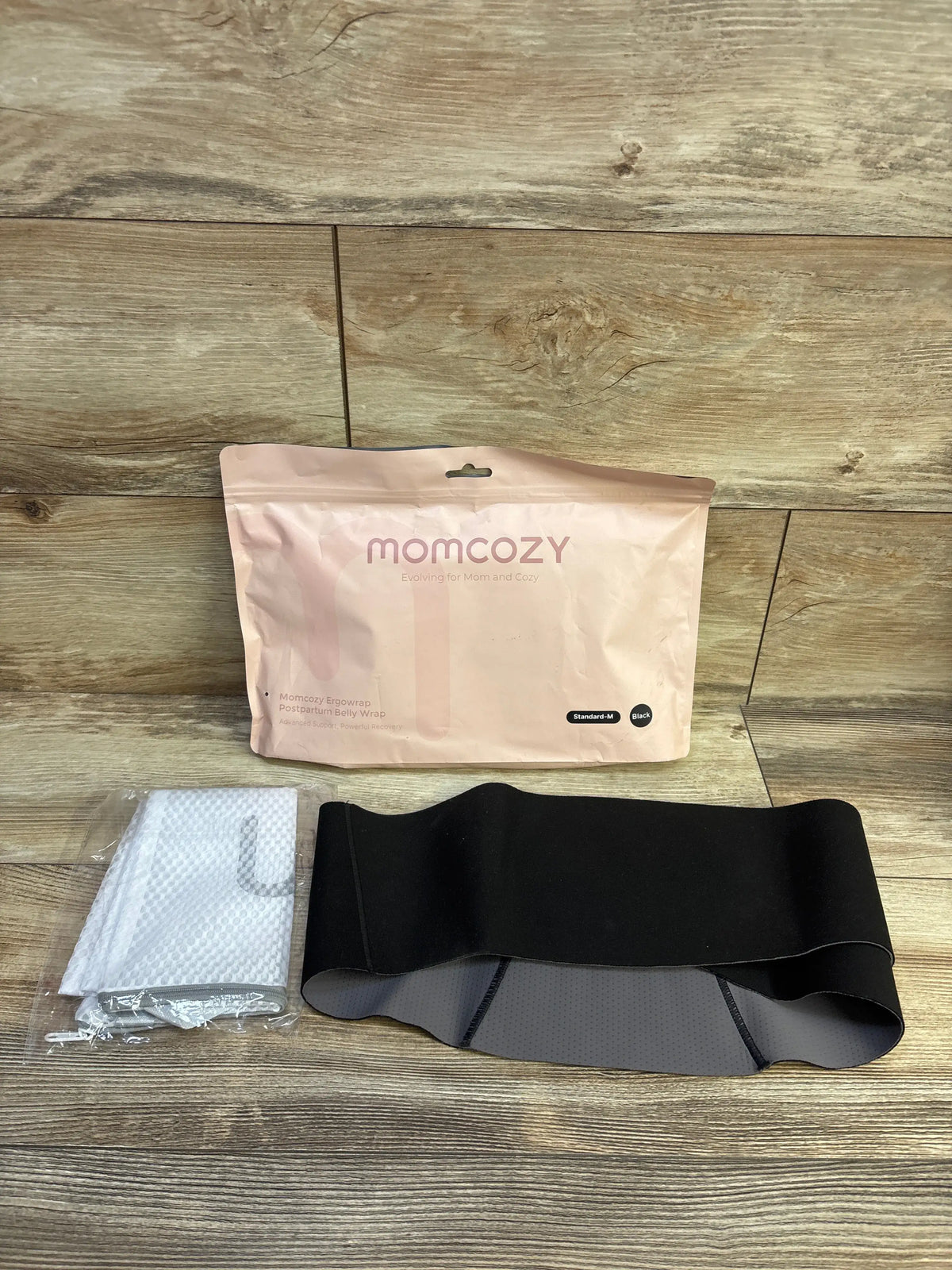 Front view of black Momcozy Ergowrap postpartum belly wrap showcasing layered support panels with packaging on a wood floor