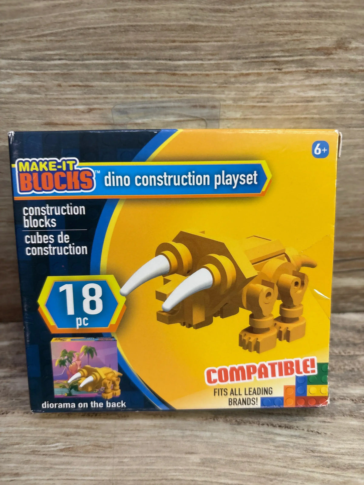 Front view of Make-It Blocks Dino Construction Playset box showing yellow triceratops build with white horns and 18 pc badge on wood background