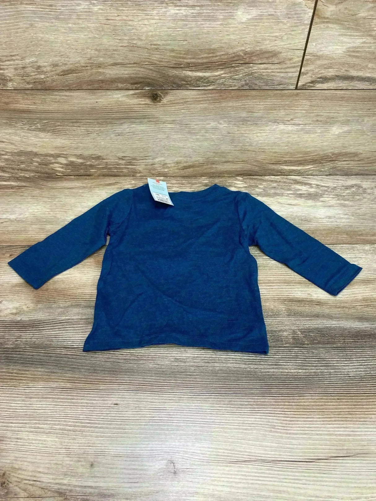 Flat lay of NEW Cat and Jack dragon shirt sz 12m showcasing solid blue long sleeve top with original hangtag on wood plank floor