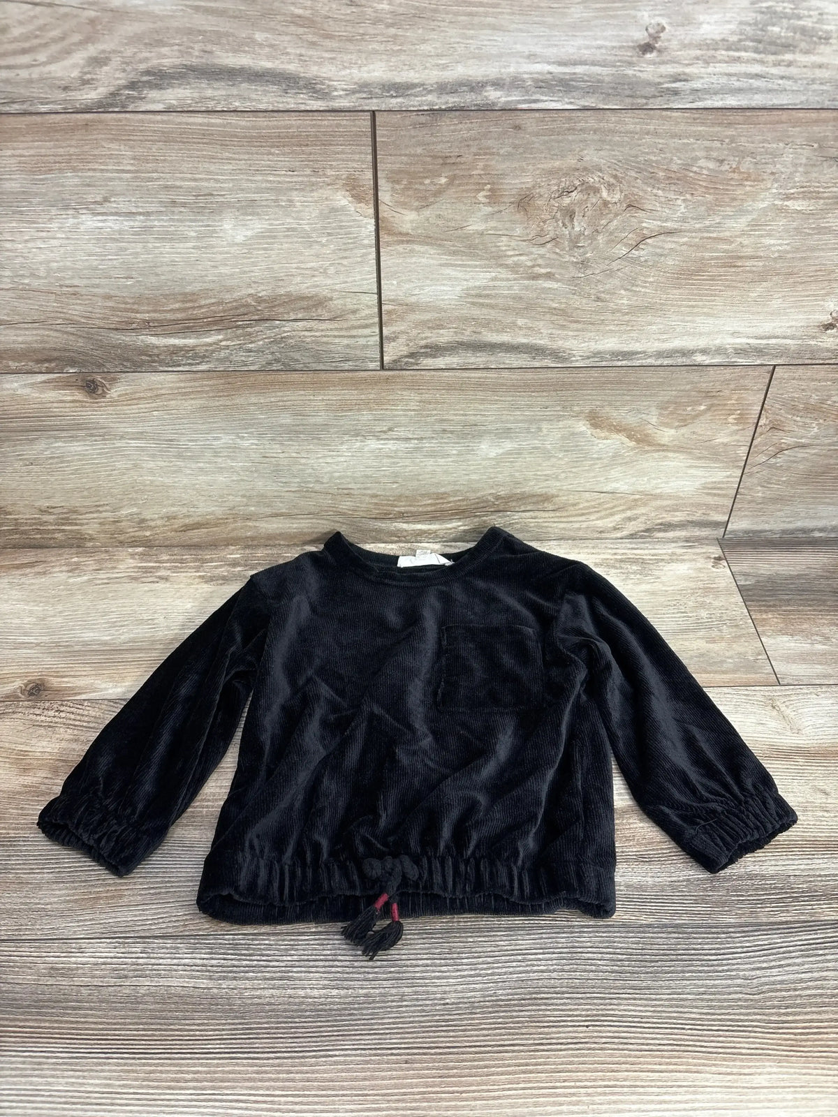 Black long sleeve toddler shirt with front pocket and drawstring hem on wood floor