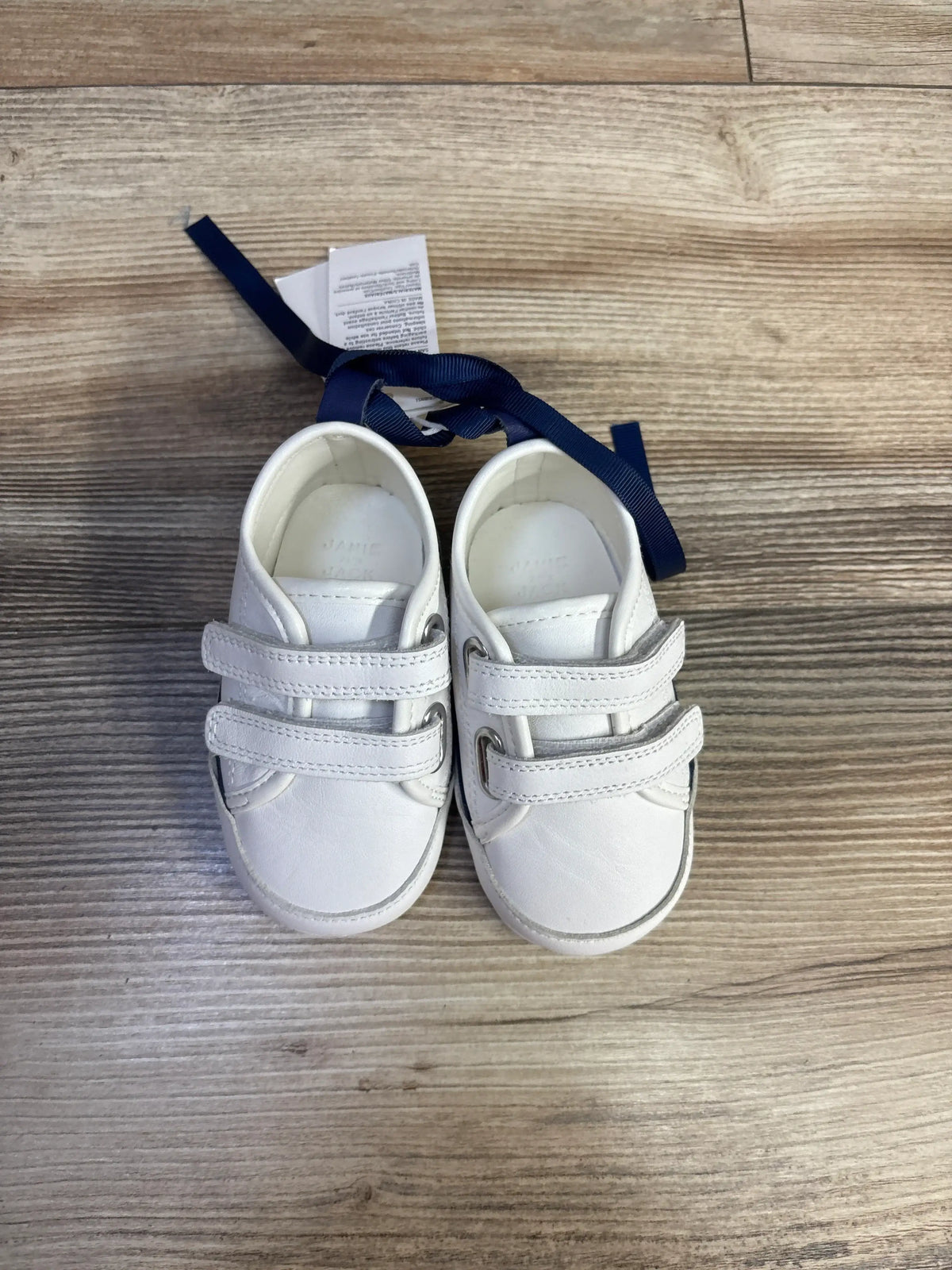 Top view of Janie and Jack double strap sneakers showcasing white uppers and two straps on each shoe on a wood look floor