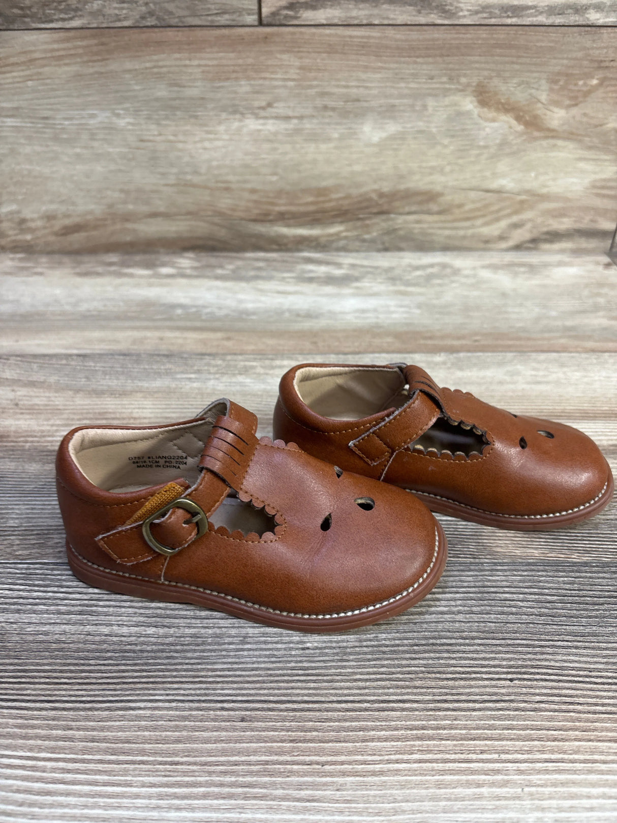 Angled front view of Otter Momo T-strap Mary Jane toddler shoes showcasing scalloped cutouts, buckle strap, and stitched sole on a wood background