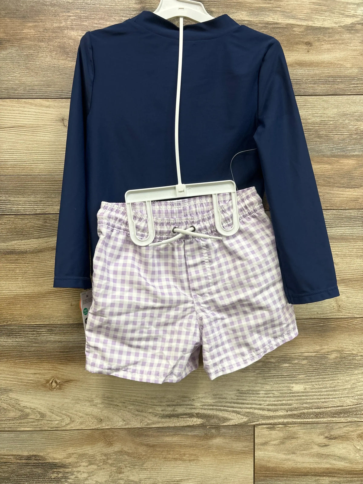 Back view of navy rash guard top and lavender gingham swim shorts on hanger against wood background
