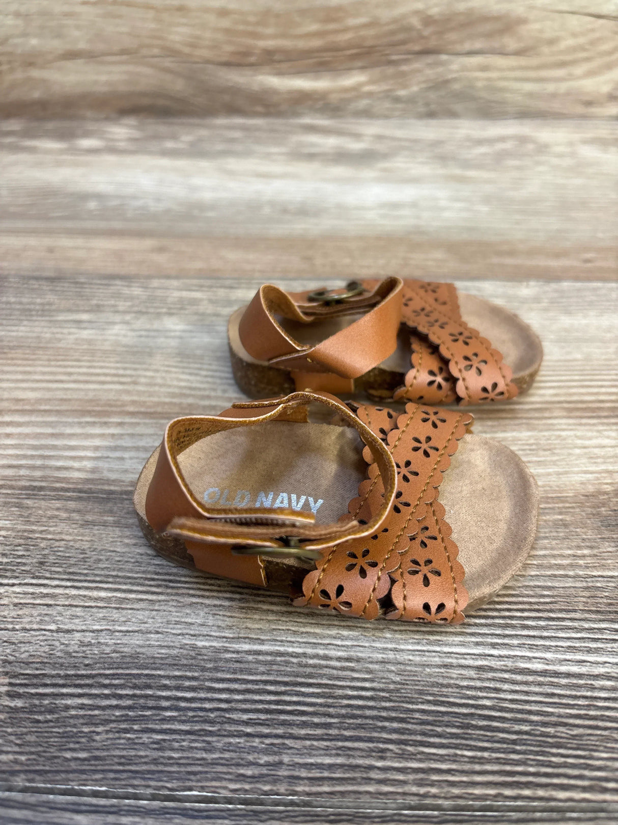Angled view of Old Navy beige faux-leather baby sandals with perforated floral straps and hook and loop closure on wood
