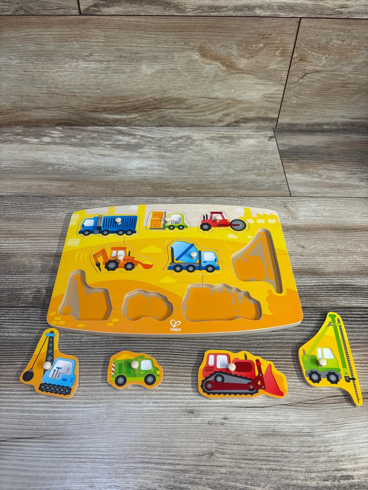 Top-down view of Janod Wooden Shapes & Sounds 6-piece block puzzle showcasing colorful vehicle wooden pieces with white knobs on a yellow board on a wood floor