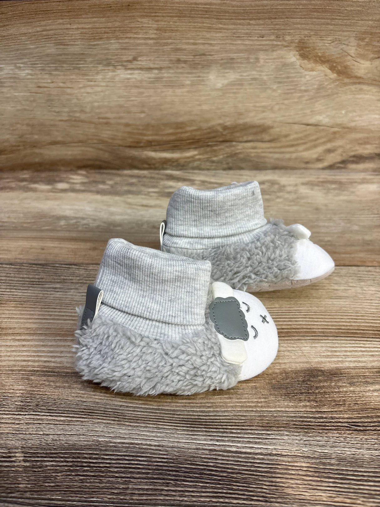 Side view of plush lamb non slip gray baby booties showing sheep face, ankle cuff and faux shearling trim on wood background