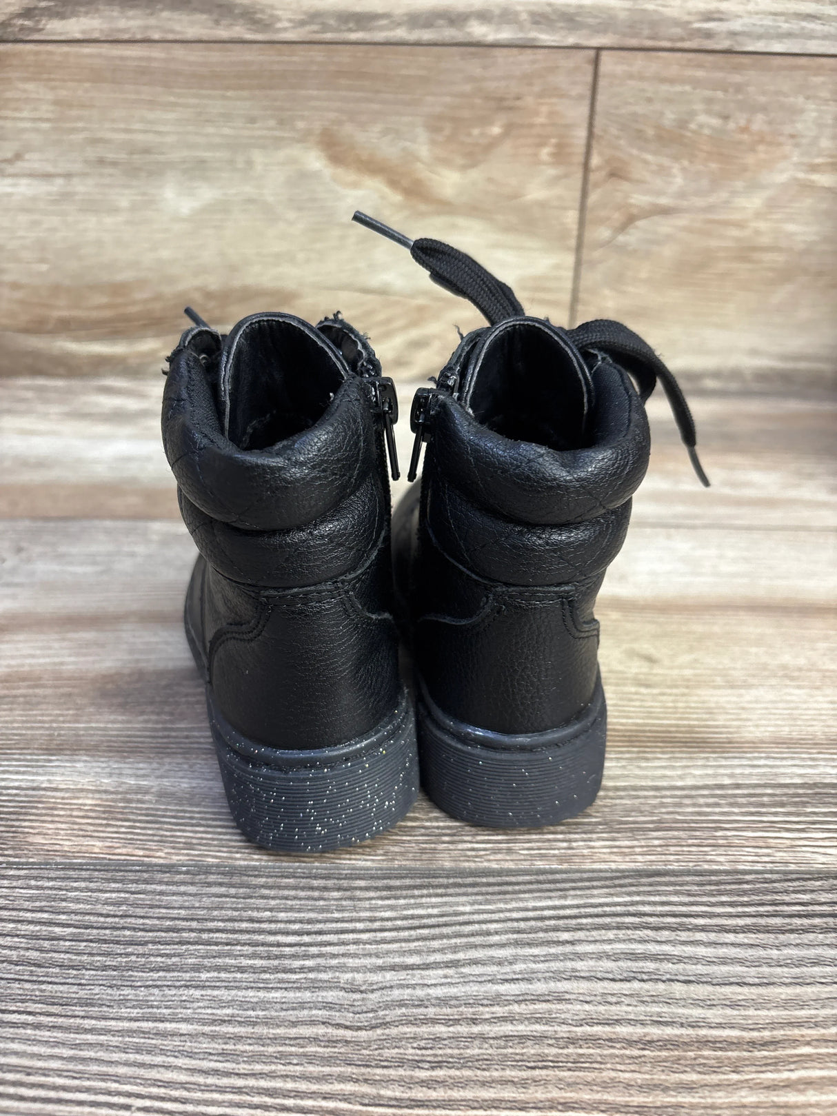 Rear view of Sincerely Jules glitter lace-up toddler boots showcasing black pebbled leather, heel zippers and glitter-speckled soles on wood background
