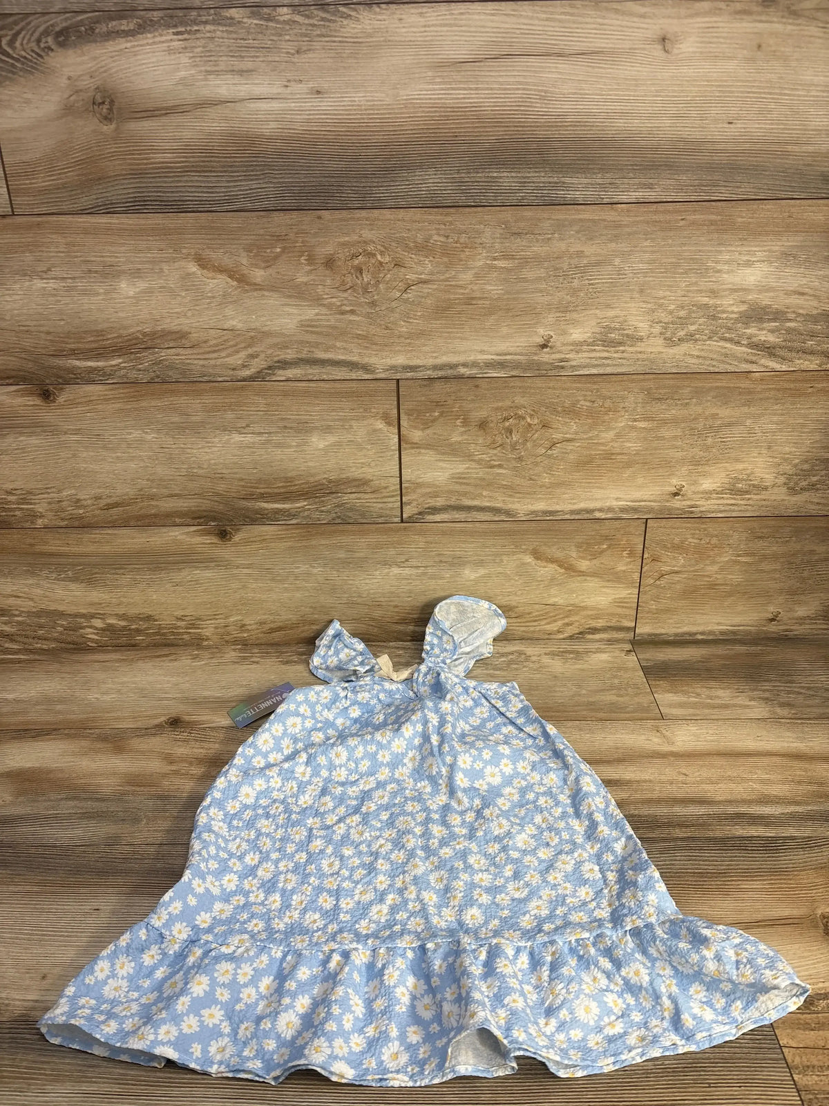 Flat lay of girls blue daisy-print sleeveless dress showcasing bow-tie shoulder straps and tiered ruffle hem with wood plank floor background