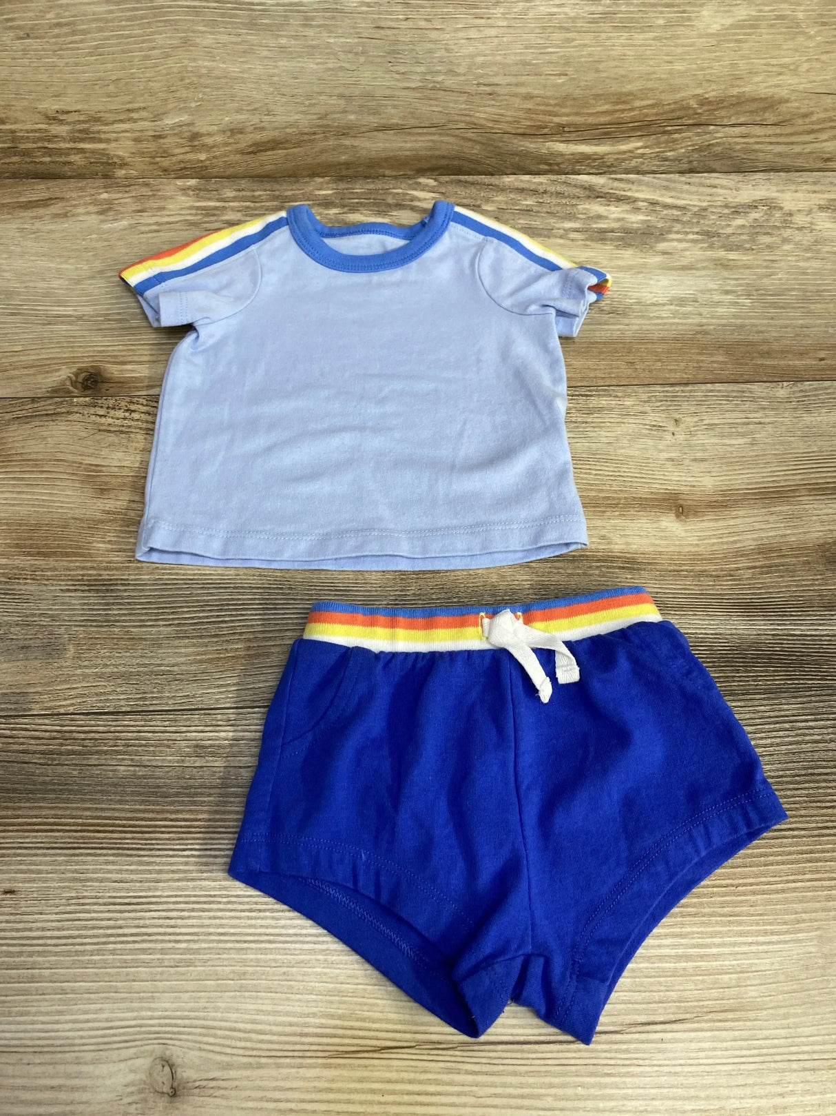 Flat lay of Cat & Jack blue baby shirt and shorts with rainbow sleeve and waistband stripes and white drawstring on wood