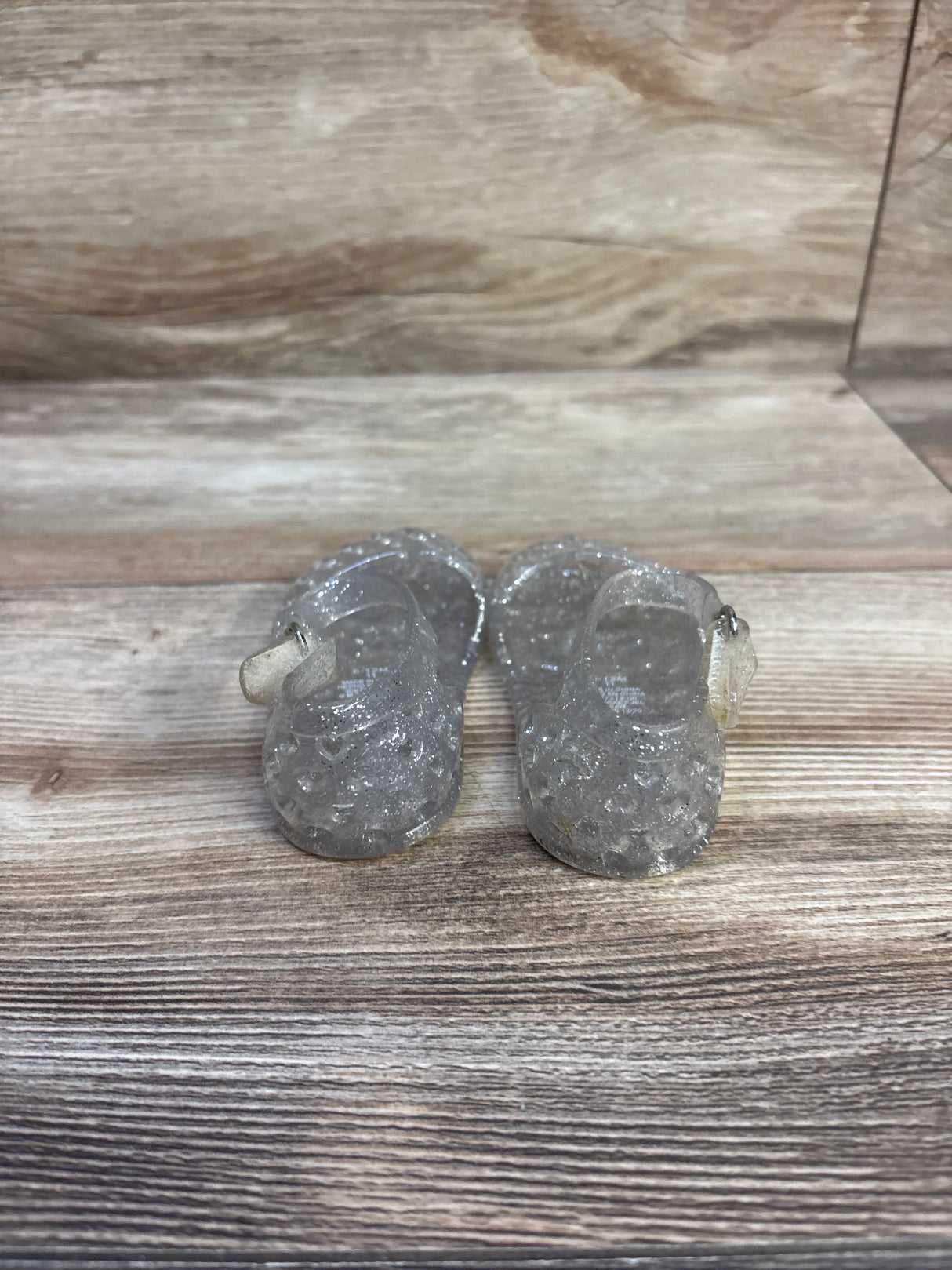 Front view of clear glitter baby jelly sandals showcasing textured toe pattern and side buckle with wood background