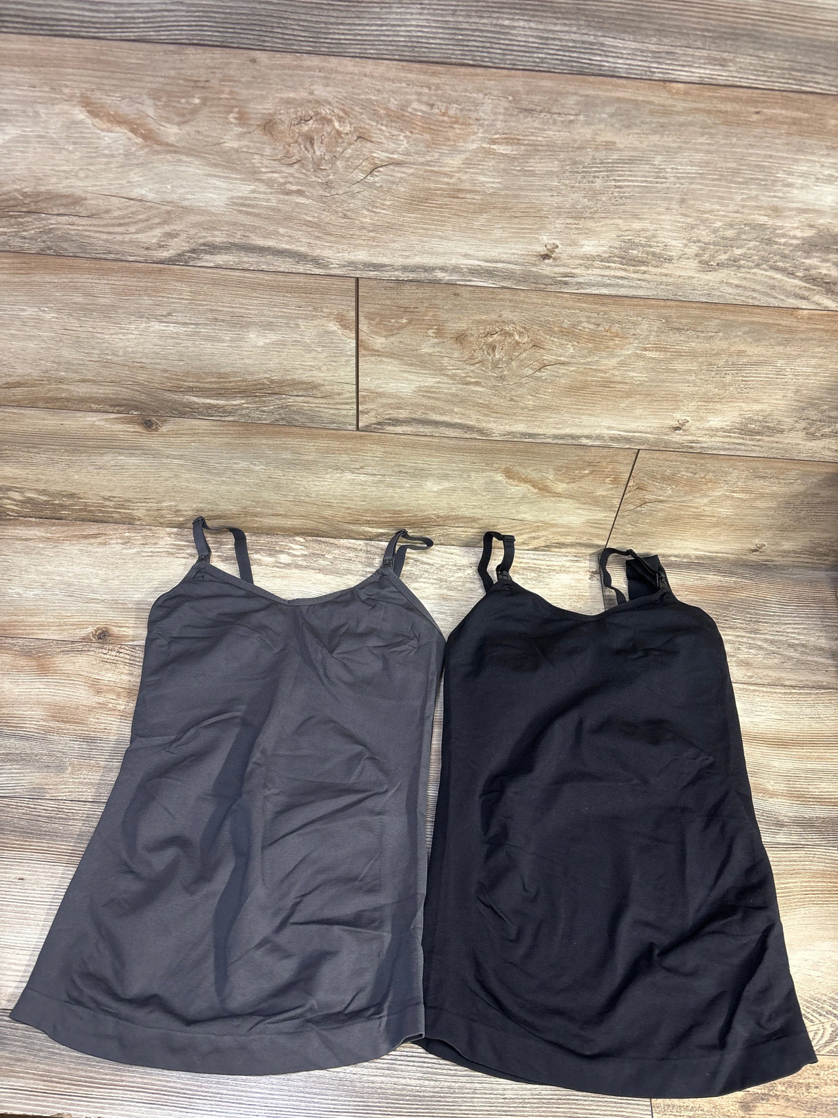 Flat lay of two nursing tank tops in grey and black showing adjustable straps and scoop necklines on wood floor