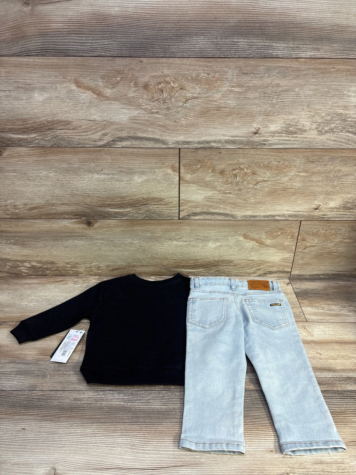 Flat lay of toddler Volcom 2-piece set showing a black long-sleeve shirt with tag and light-wash jeans with back pockets on wood background