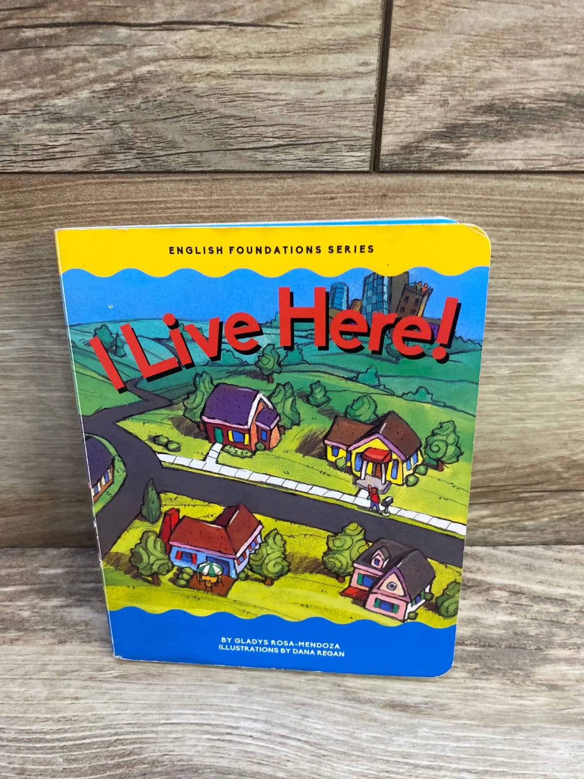 Front view of I Live Here! board book showing illustrated neighborhood cover with large red title I Live Here! on wood