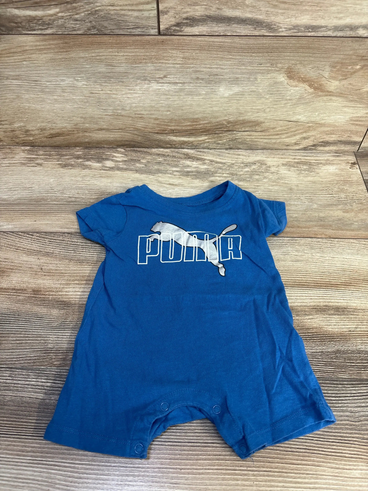 Front view of a blue 100% cotton Puma shortie romper showcasing the white Puma logo and snap closures against a wood-grain background