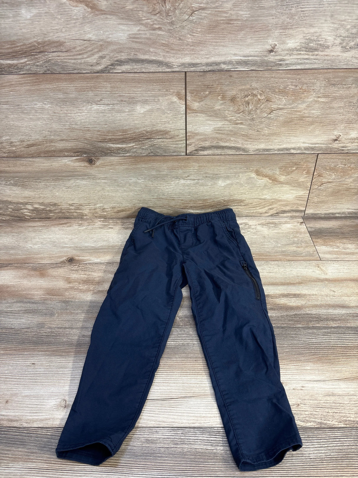 Flat lay of boys’ navy tech taper quick-dry pants showcasing drawstring elastic waist and zippered side pocket on wood floor