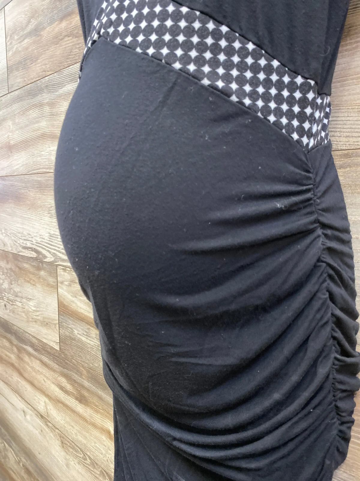 Side view of Kimi + Kai black maternity bodycon dress showing ruched side and patterned waistband on wood background