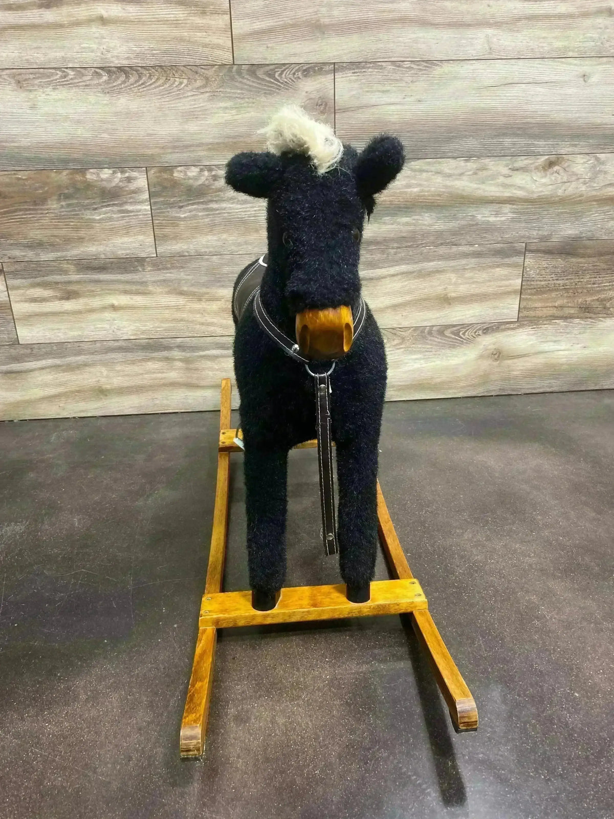 Front view of vintage black plush rocking horse with light mane, wooden rocker base and leather bridle against wood-paneled background